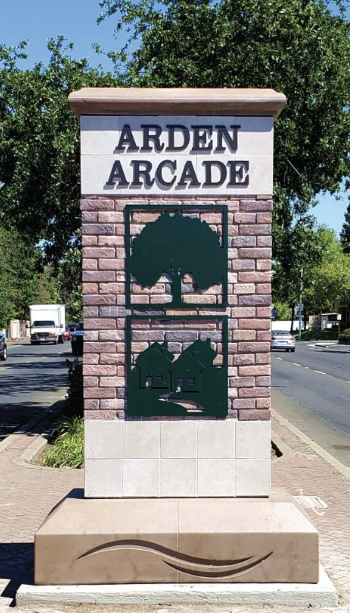 Sign reading "Arden Arcade" with tree and houses design, set on a brick structure in the median a road with trees.