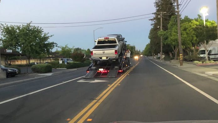 A tow truck is transporting a pickup truck along a street, with trees and buildings visible on either side.