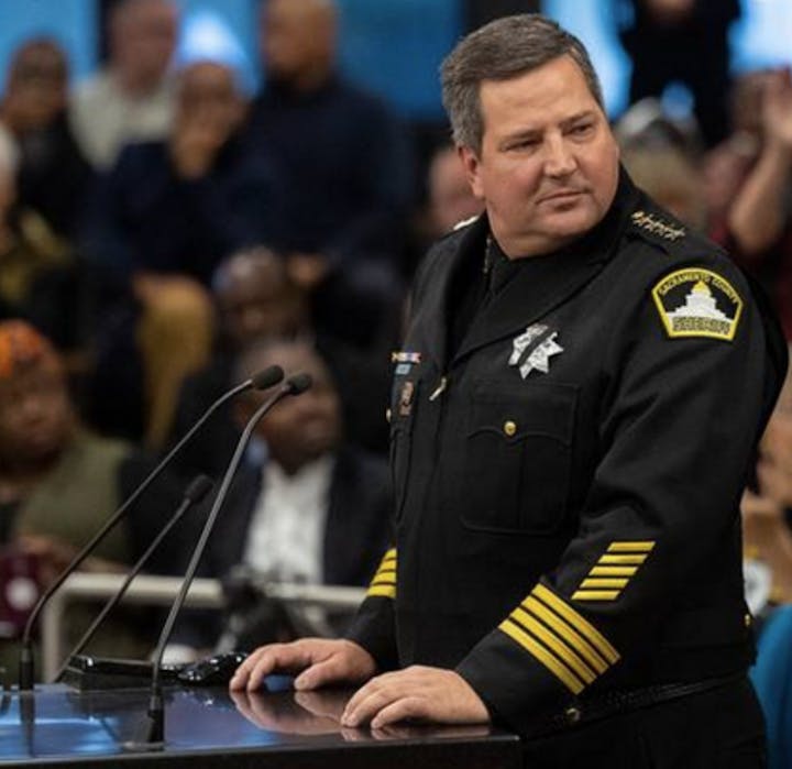 A police officer in uniform stands at a podium, addressing an audience with people in the background.