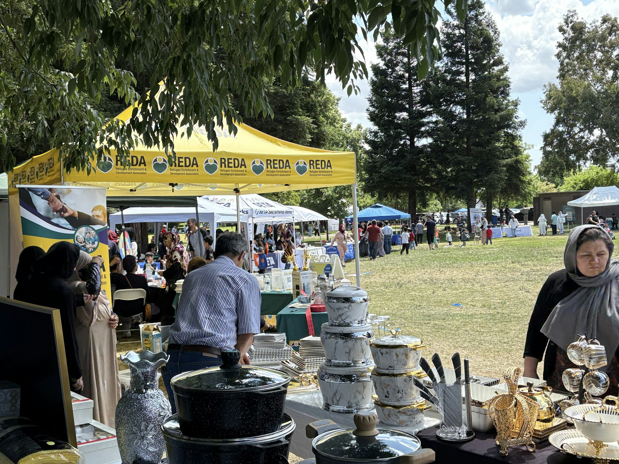 An outdoor market with people, various stalls with crockery, cutlery, and a RECA-branded yellow tent.