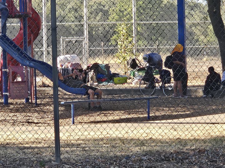 People sitting and standing near a playground slide, with bags and a bicycle nearby, seen through a chain-link fence.