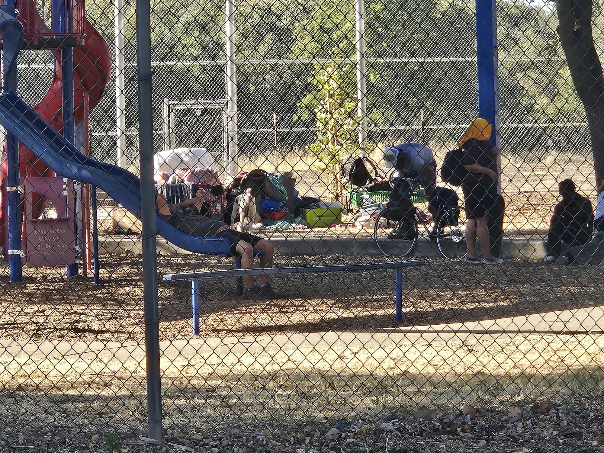People sitting and standing near a playground slide, with bags and a bicycle nearby, seen through a chain-link fence.