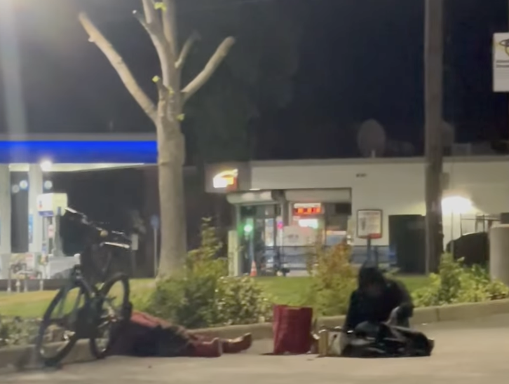 The image shows two people at night, one lying down and another sitting nearby, with a bike and a gas station in the background.