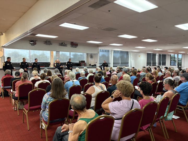 A public meeting with police officers speaking to a large audience seated in a hall.