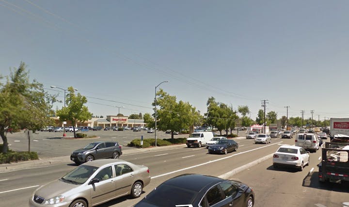 A busy street scene with parked cars, trees, and a shopping plaza in the background under a clear blue sky.