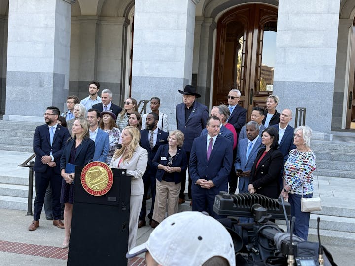 A group of people standing on steps outside a building, with a podium displaying the California State Senate seal.
