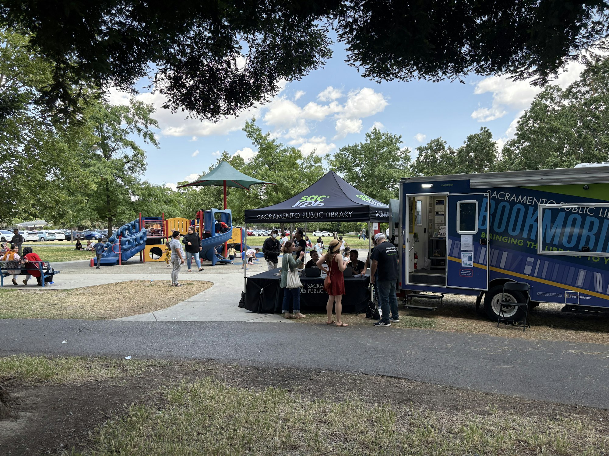 Library bookmobile and pop-up tent with people and with children playing at the adjacent play structure.