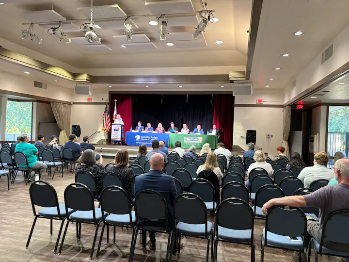 A meeting or panel discussion is taking place with a small audience present, featuring a speaker and several panelists at tables.