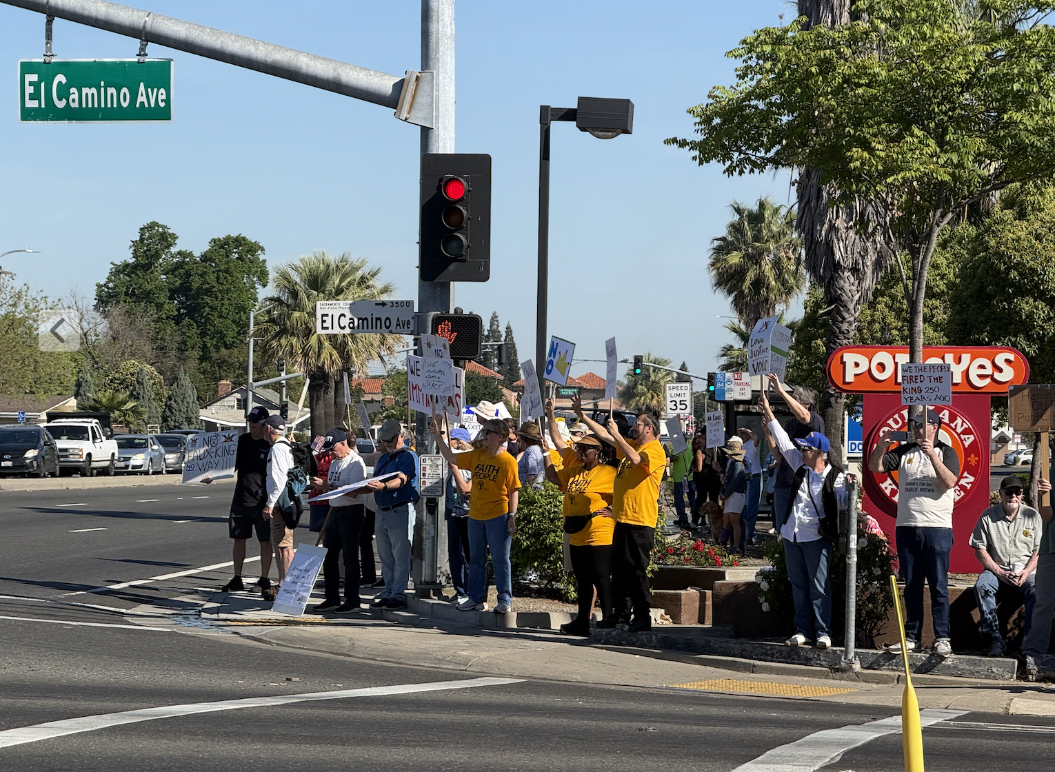 A group of protesters holding signs at a busy intersection on El Camino Ave, with a Popeyes restaurant in the background.