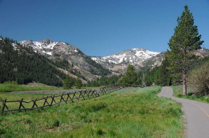 A scenic landscape featuring rolling hills, snow-capped mountains, a paved path, and lush greenery under a clear blue sky.