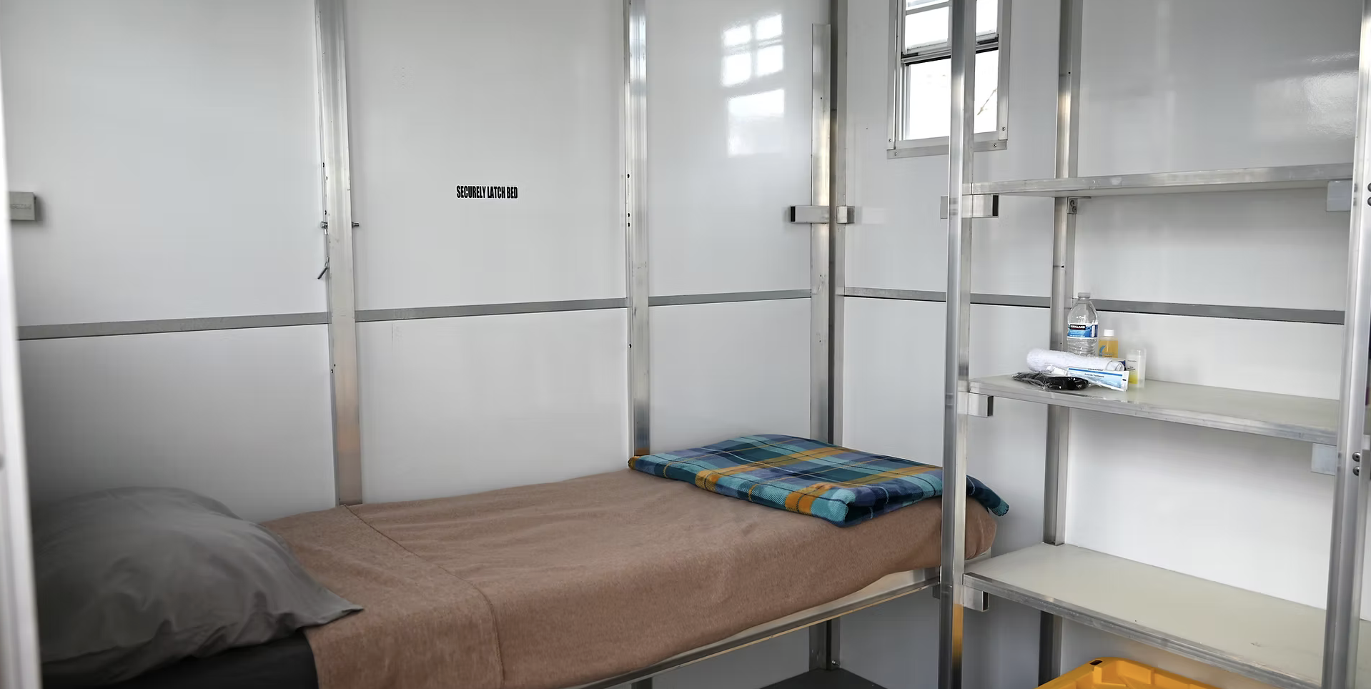 A small room with a bed, plaid blanket, shelves, a water bottle, toiletries, and a "Securely Latch Bed" sign.