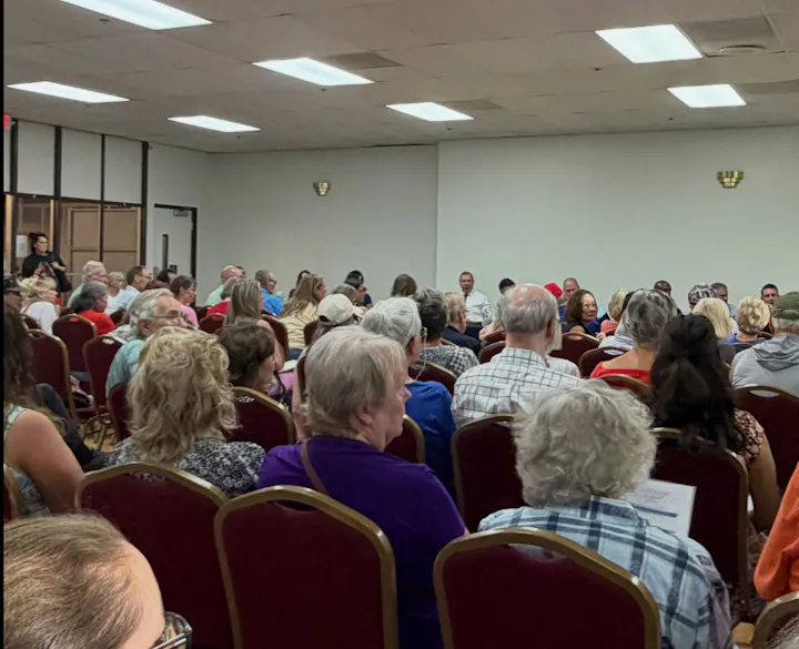 A large audience is seated in a room, likely attending a meeting or event, with various people visible in multiple rows.