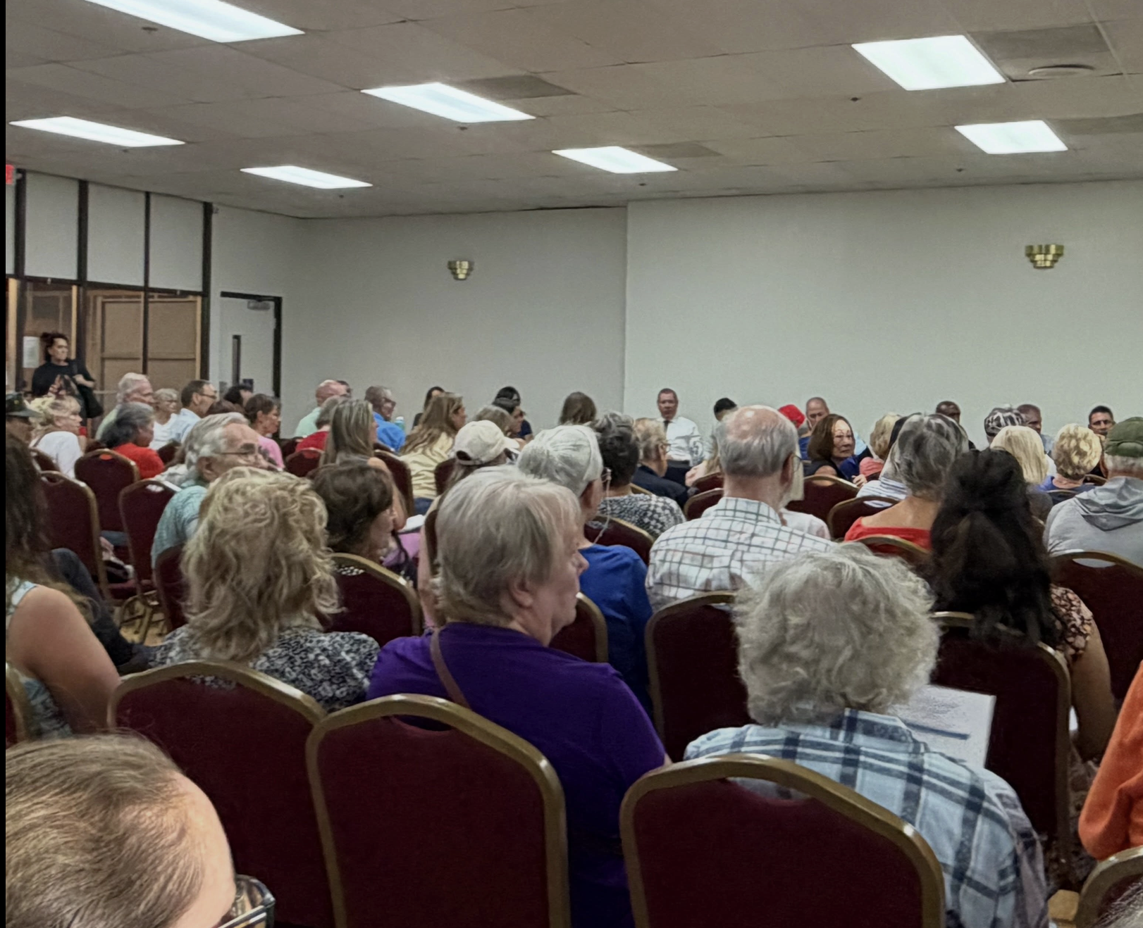 A large audience is seated in a room, likely attending a meeting or event, with various people visible in multiple rows.