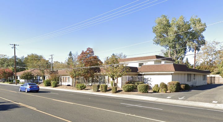 A blue car on a road, beside a white building with brown roof, surrounded by trees and power lines under a clear sky.