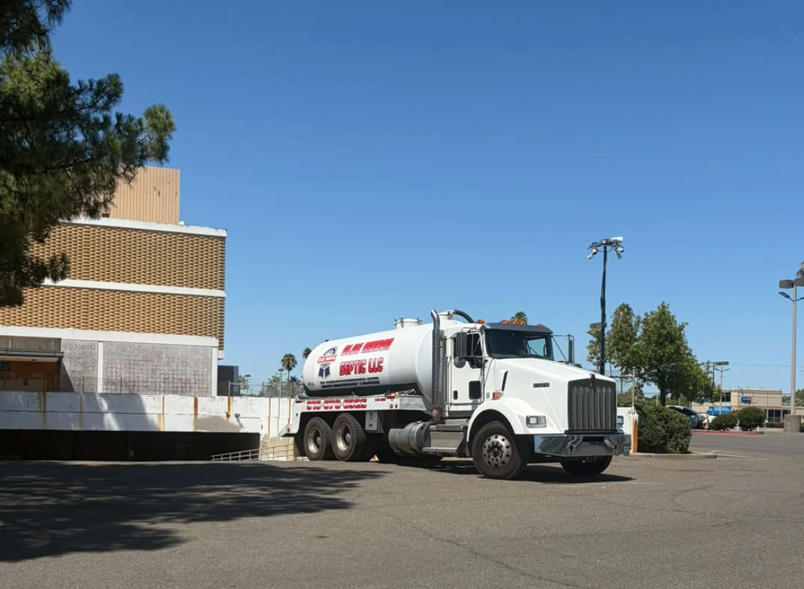 A white septic truck is parked near a building on a clear day, with trees and empty parking spaces around.
