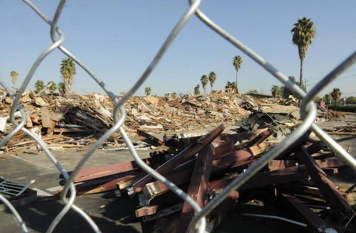 A view through a fence of a construction site with rubble and debris, palm trees in the background under a clear blue sky.