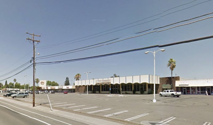 A commercial plaza with several stores and a large empty parking lot, featuring palm trees and power lines against a clear sky.