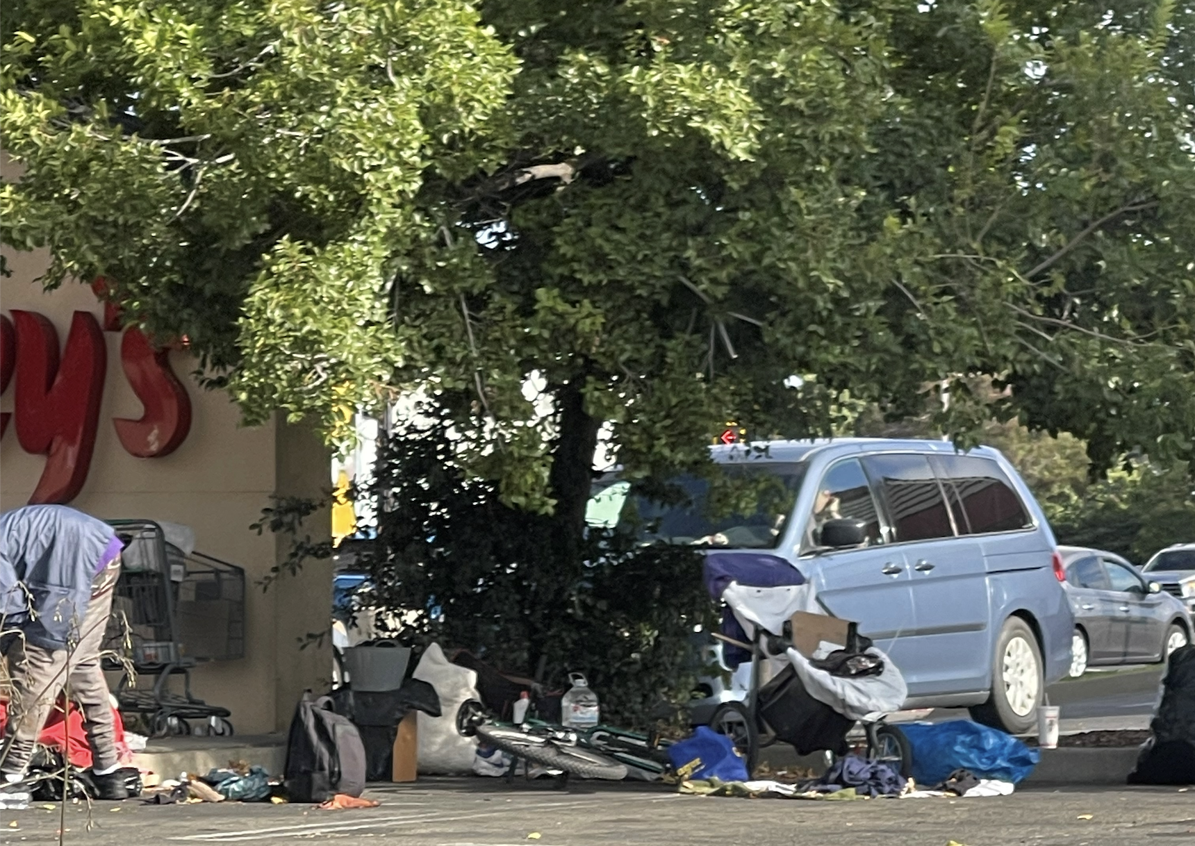 A person stands near scattered belongings and shopping carts by a building, with a blue vehicle parked nearby.