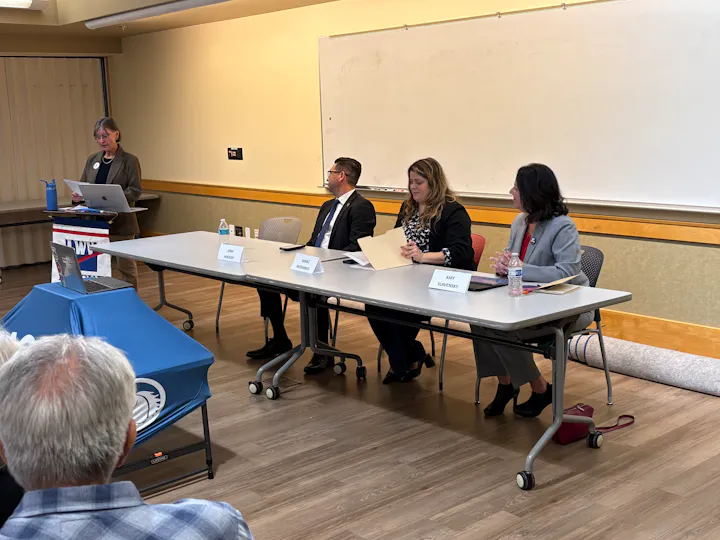 A speaker addresses an audience while four panelists sit at a table, engaging in a discussion or presentation.