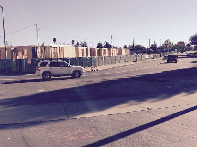 A road with vehicles passing by, and a construction site with wooden frames and a green fence in the background.