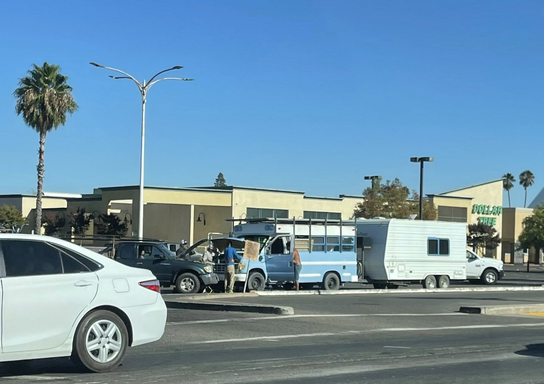 Vehicles parked in a lot, including a blue bus and white camper, with a person holding a sign near a store.