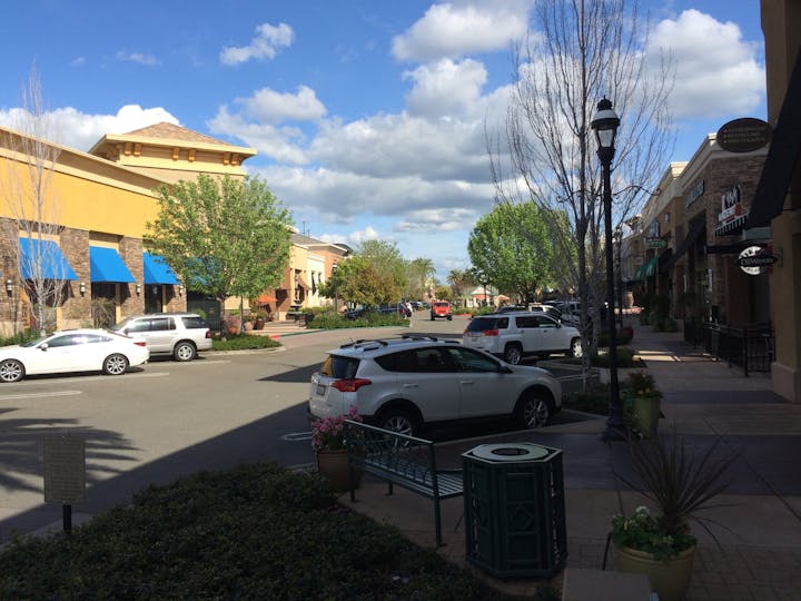 A bustling shopping area with greenery, storefronts, and parked cars under a partly cloudy sky.