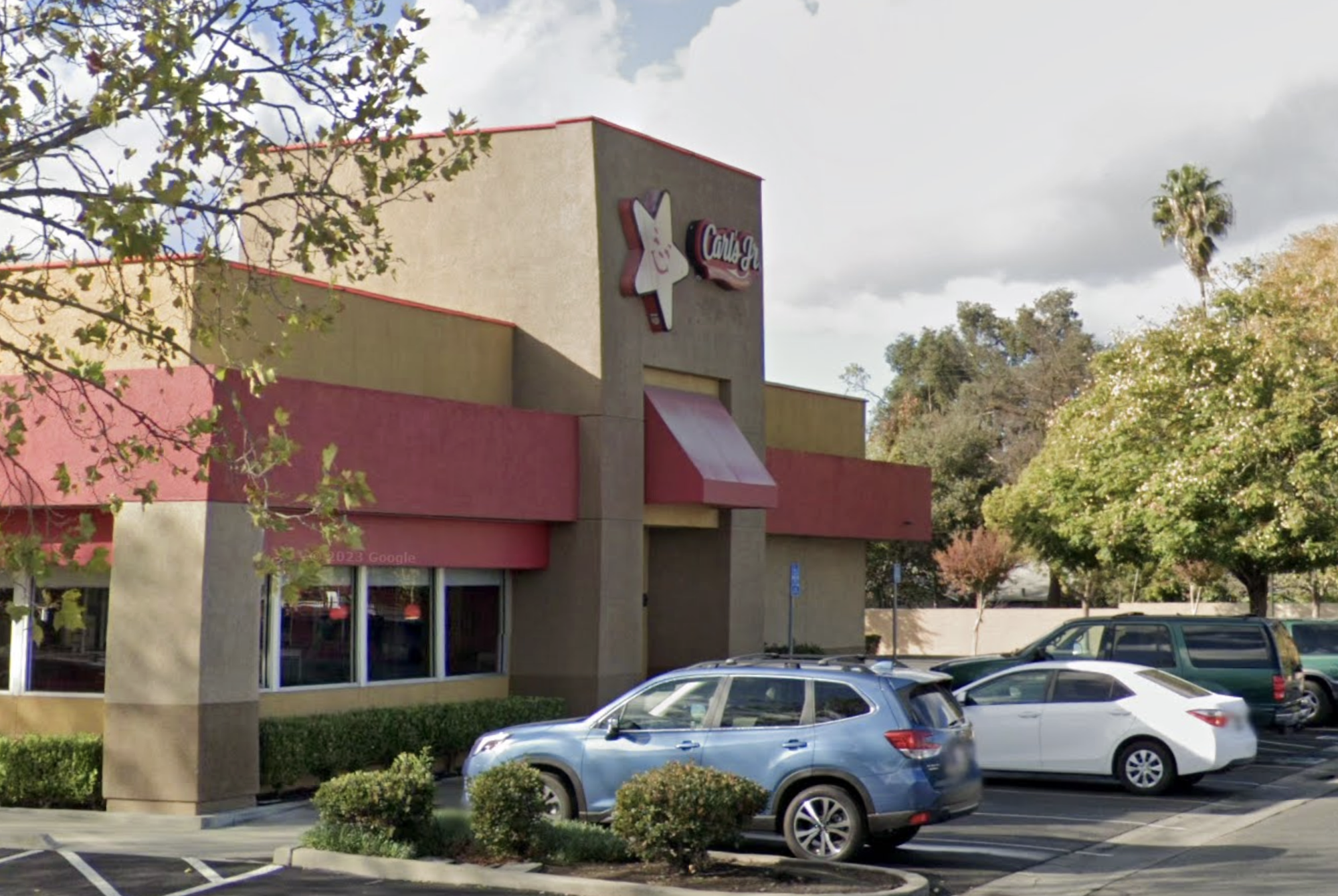 A fast-food restaurant with a red awning and cars parked outside, surrounded by trees.