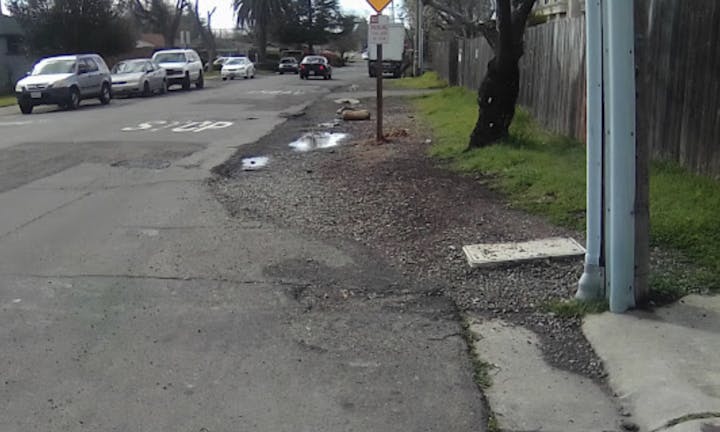 The image shows a street with parked cars, a stop sign, some puddles, and gravel along the roadside.