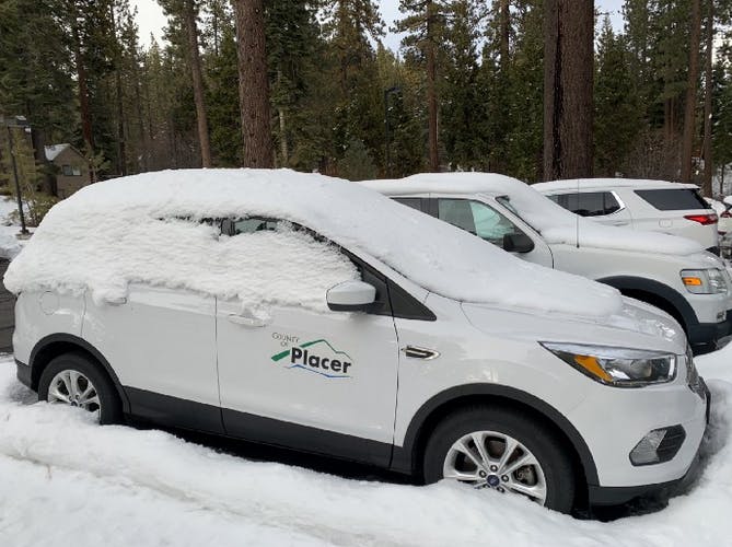 A white vehicle covered in snow, parked among trees, with a "County Placer" logo visible.