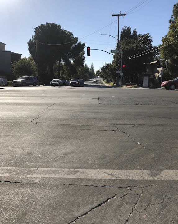 The image shows a sunlit road intersection with a red traffic light, cracked pavement, and trees lining the street.