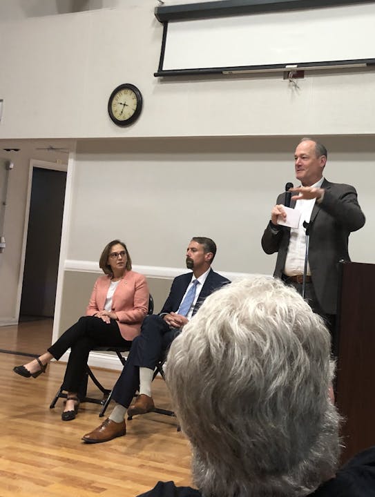 A speaker with a microphone stands addressing an audience; two individuals sit behind him, suggesting a panel discussion or presentation.