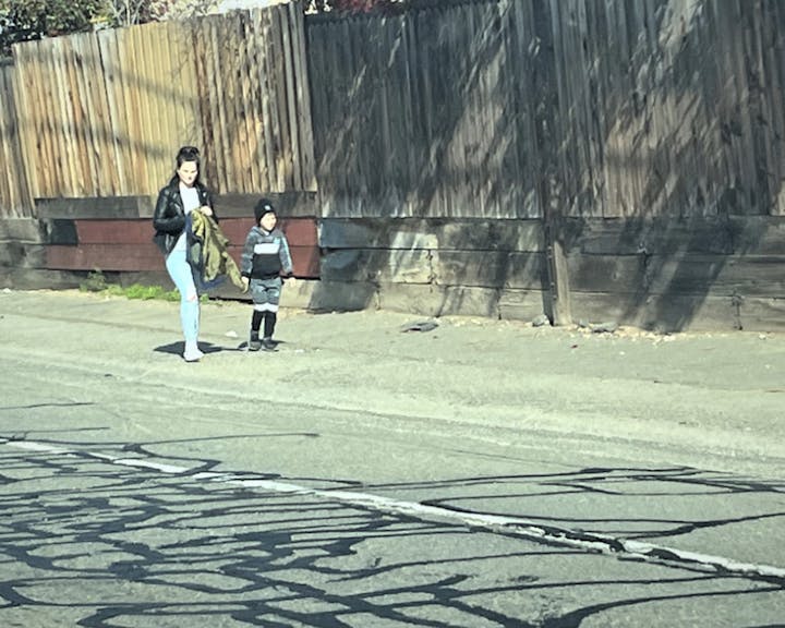 An adult and a child walking by a wooden fence, with cracked street pavement in the foreground.