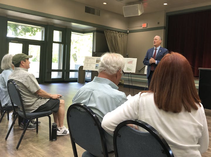 A man speaks to seated listeners in a room with informational posters on easels.