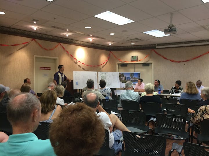 A community meeting is taking place with attendees listening to a speaker and viewing presentation materials on a board.