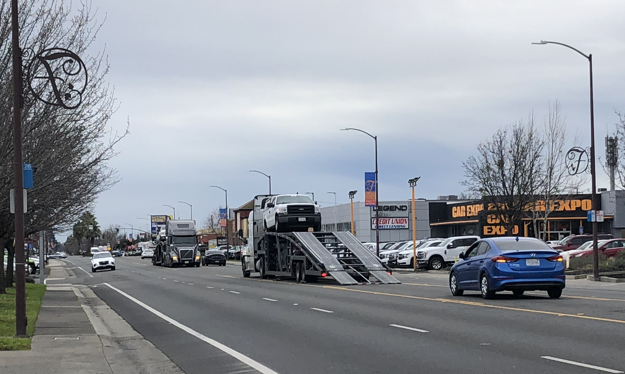 Urban street with cars, a car hauler truck, and a "CAR EXPO" sign.