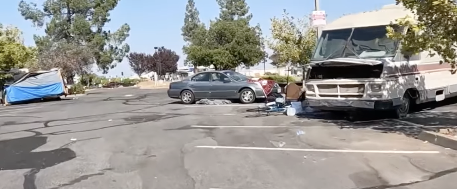 Parking lot scene with a car, RV, bicycle, various items on the ground, trees in the background.