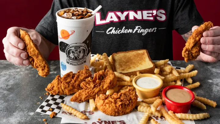 A meal featuring chicken fingers, French fries, toast, dipping sauces, and a soda, with a person holding a chicken finger.