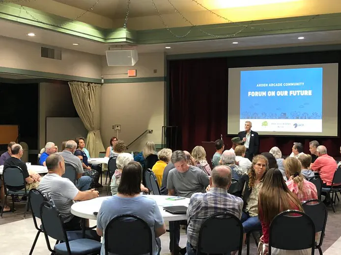 A community forum in Arden Arcade, with attendees seated at tables, discussing future plans led by a speaker.