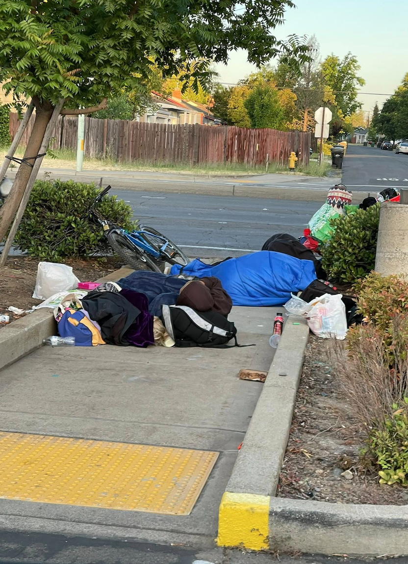 A person sleeping on a sidewalk with belongings and sleeping bags, a bicycle, and a nearby street with trees and houses in the background.
