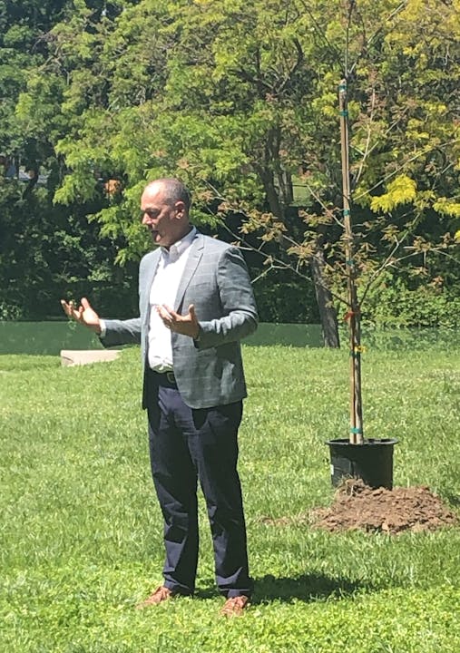 A man gesturing in a park with a young tree and soil behind him.