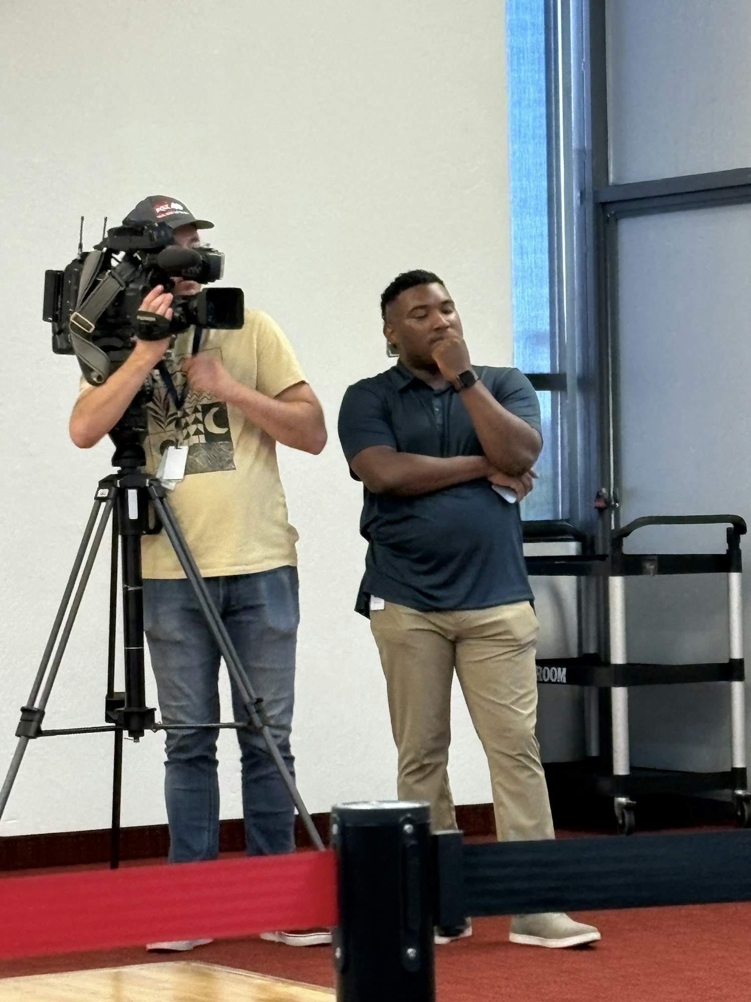 A news person stands next to a person operating a video camera on a tripod.