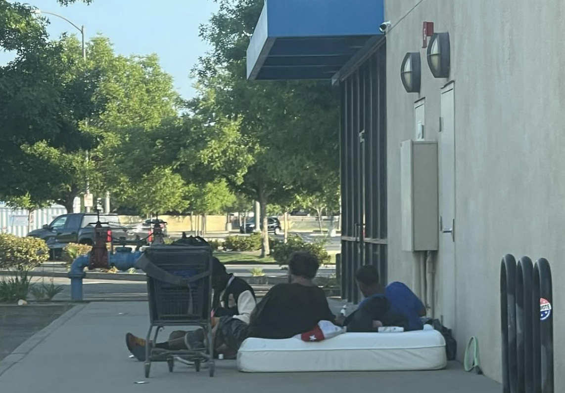 People resting on a mattress on a sidewalk next to a building, with a shopping cart nearby.