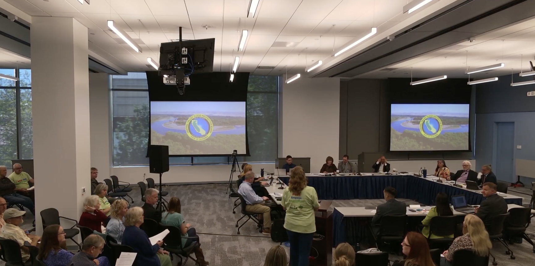 A meeting room with people seated; two screens display a logo and aerial image.
