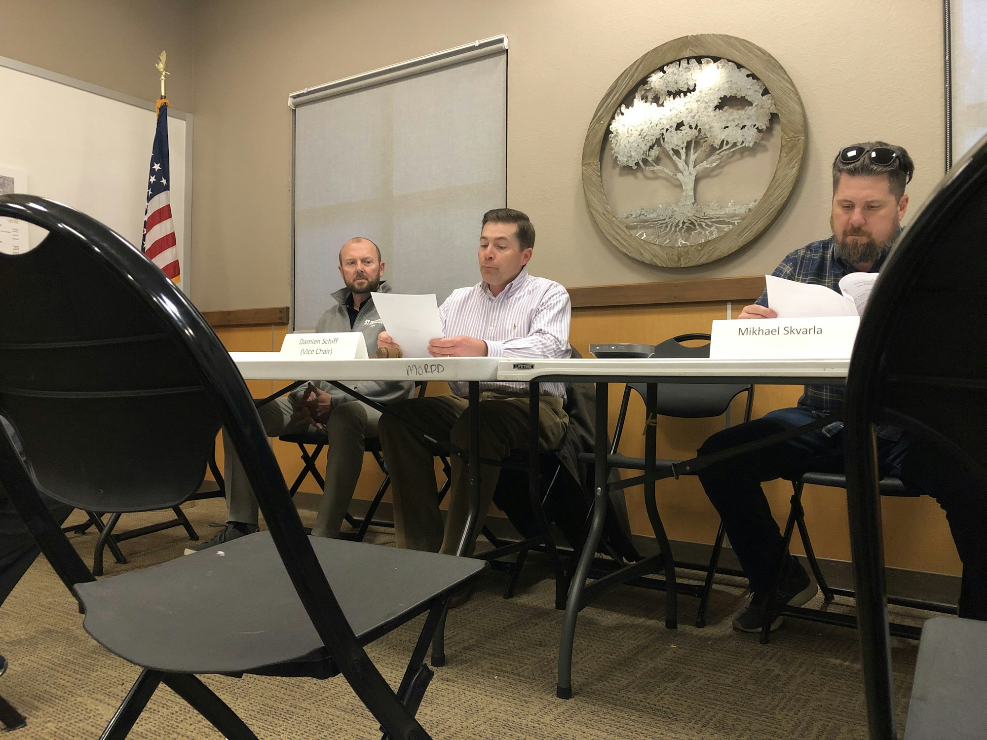 Three men sitting at a table with name placards, one reading a paper, in a meeting room with a US flag and a wall art.