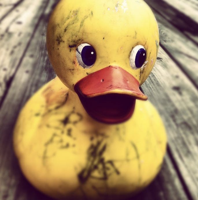 The image depicts a large, weathered yellow rubber duck with a bright orange beak, sitting on a wooden surface.