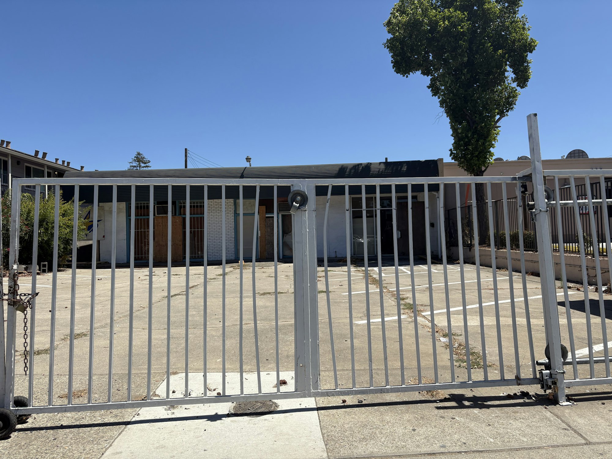 A gated, vacant parking lot with a building in the background and a tree to the right under a clear blue sky.