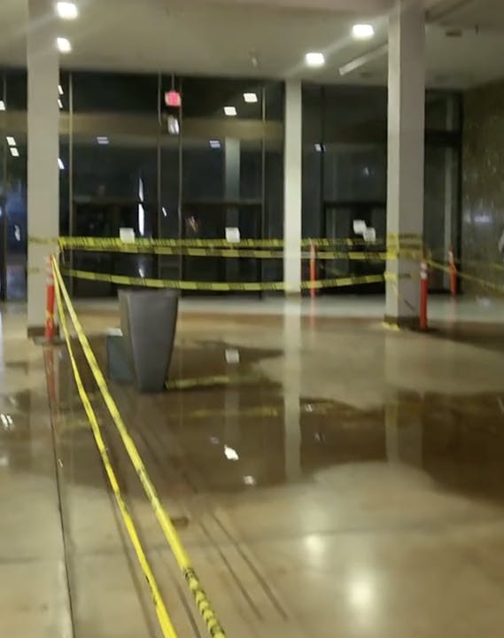 Indoor area with caution tape, wet floor signs, and a small puddle on a shiny floor, likely indicating a safety hazard.