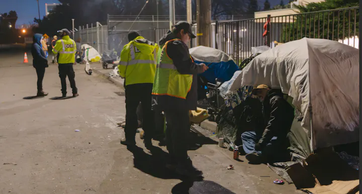 The image shows a group of people in reflective vests interacting with individuals near makeshift tents in an urban setting.