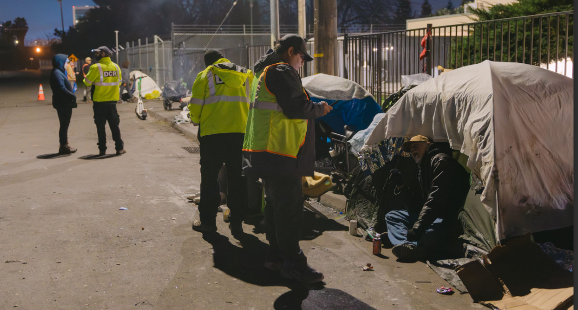 The image shows a group of people in reflective vests interacting with individuals near makeshift tents in an urban setting.