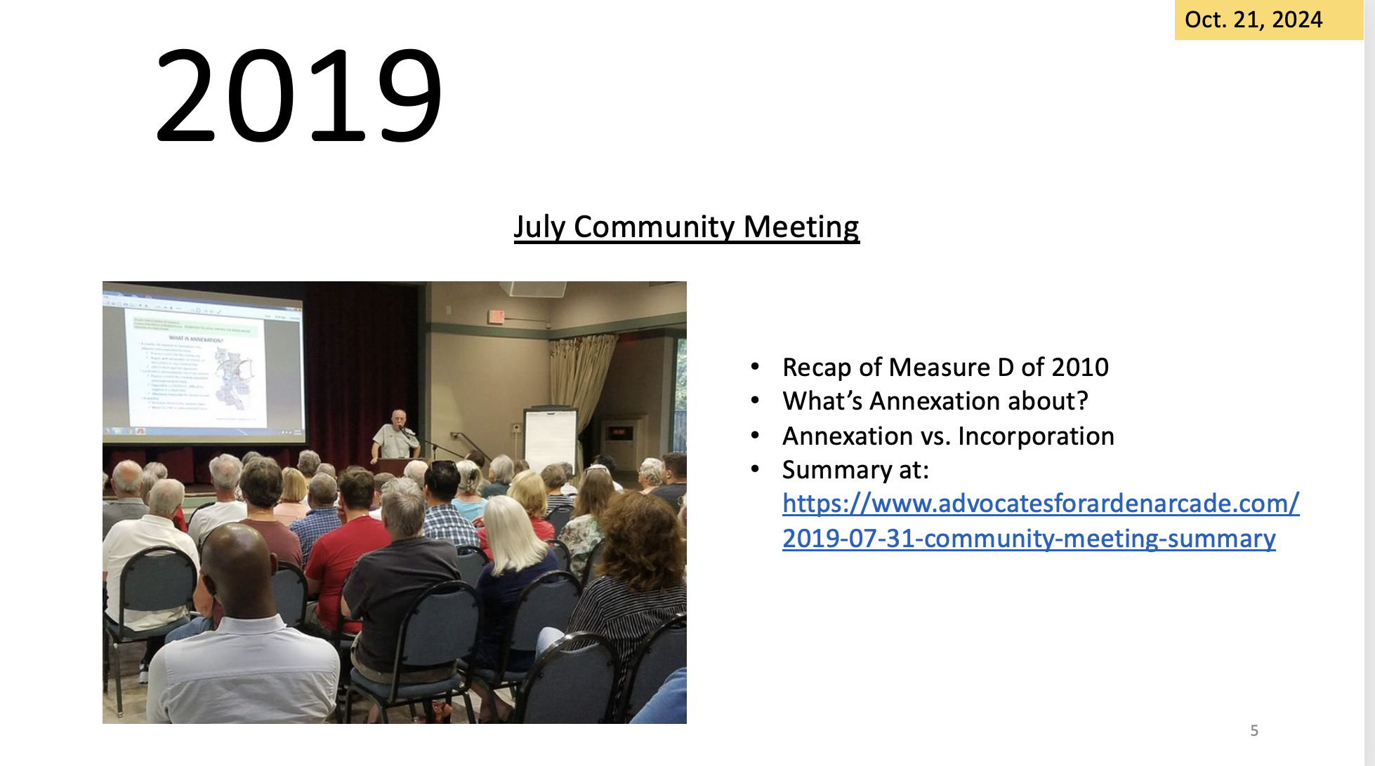 Community meeting with a presentation, attendees listening to a speaker, text and dates on a projected screen.